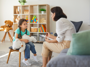 A stock photo of a child talking to a therapist in a bright room with toys on a shelf in the background.
