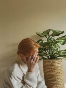 A stock photo showing a white woman sitting next to a potted plant with her head in her hands.
