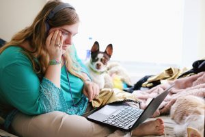 A stock photo that shows a white woman sitting on a bed with her computer on her lap, sitting next to a small dog with its ears perked up. The woman looks stressed. 