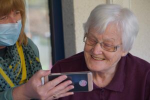 A stock photo showing an elderly white woman looking at a phone that's being held up by a younger white woman wearing a mask next to the older woman.