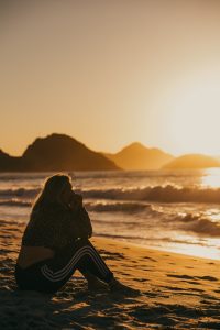 A stock photo of a woman sitting on the beach at sunset staring at the water.