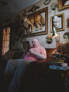 an elderly man looking at a photo frame as he sits on the couch. Above him is a taxidermy fox.