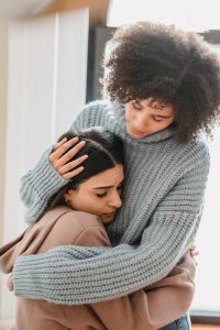 Woman with curly hair in a big gray sweater consoles another woman with black hair wearing a peach hoodie.