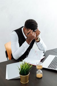 A stock photo that shows a Black man in sitting at a work desk with his head in his hands like he's overwhelmed. 